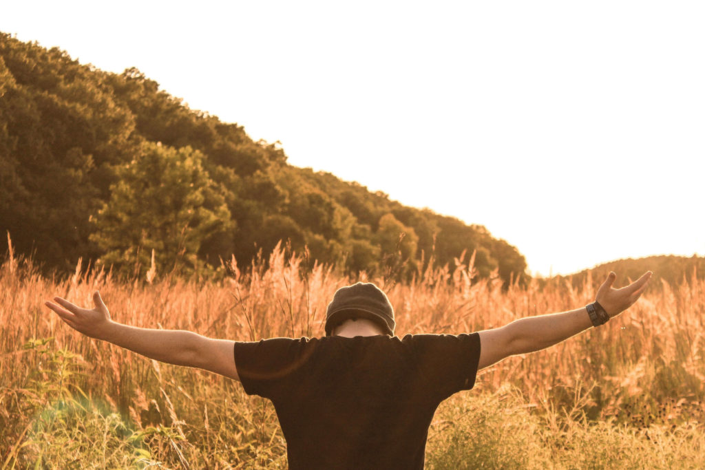 A man with a beanie on and head down back facing the camera and arms stretched wide. The field is fall and gold colors are all around