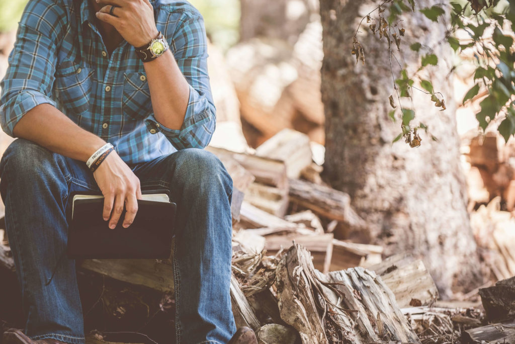 A man in denim jeans sits on logs while holding the bible. His face is not shown