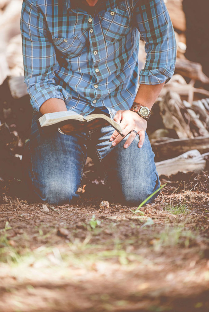 A man in denim jeans on his knees reading the bible