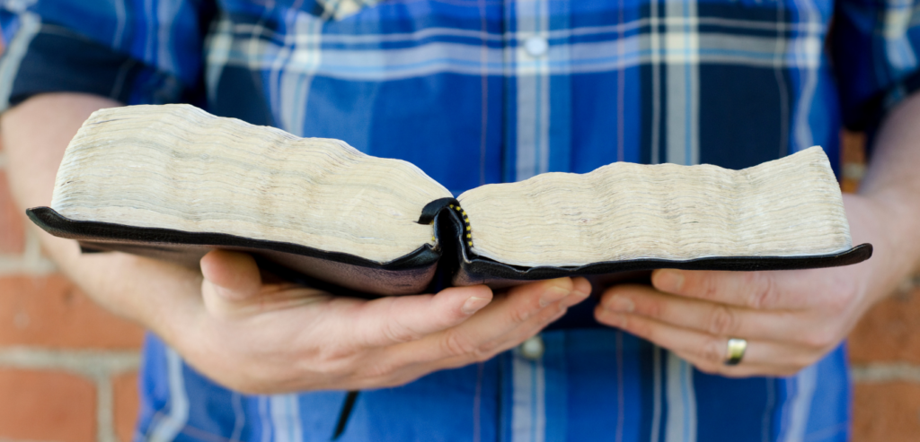 A white man in a blue plaid shirt holds a bible. Only his hands are visible, he wears a wedding ring