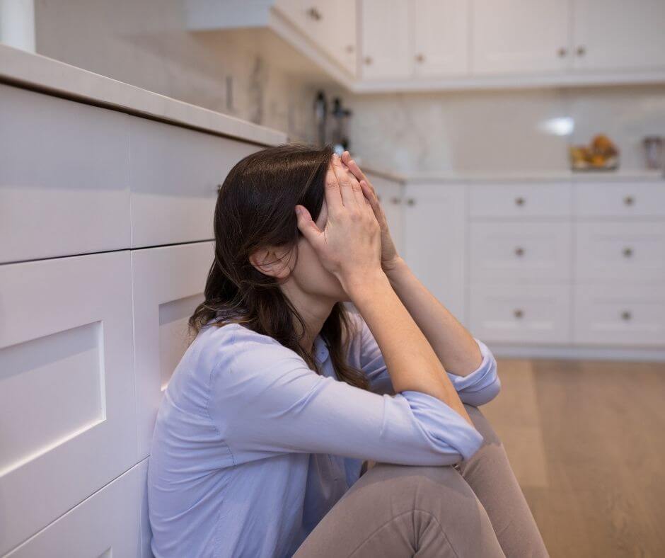 A woman sitting on the kitchen floor looking distraught with her head in her hands