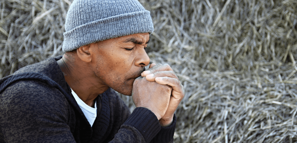 A black man sits outside, he has a stocking cap on and seems to be praying
