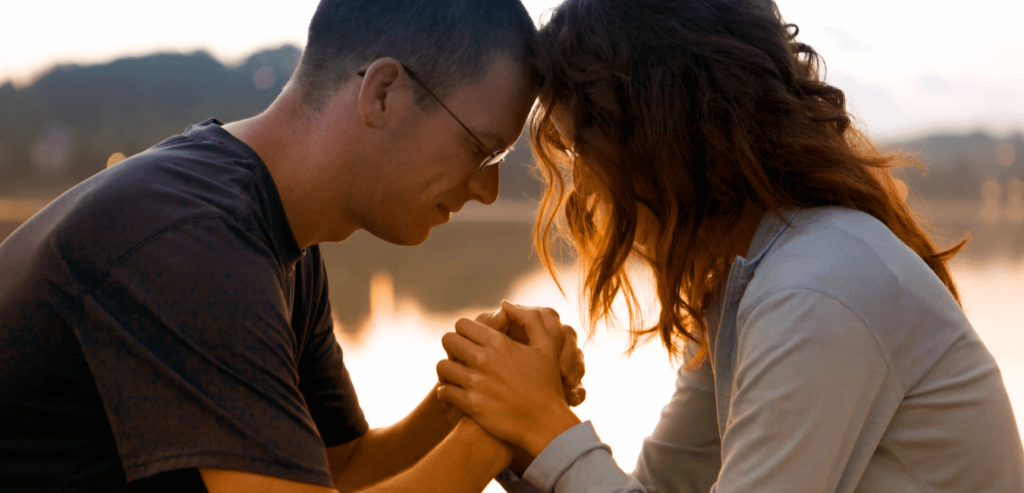 A couple who are sitting while holding each other hands and praying together at sunrise by lake