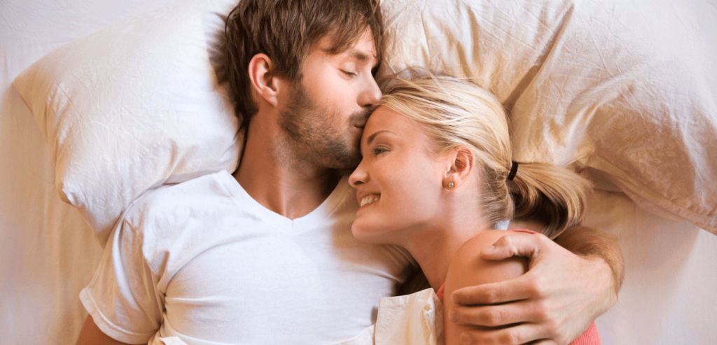 A male embracing and kissing his female partner in her forehead while they are lying on a bed