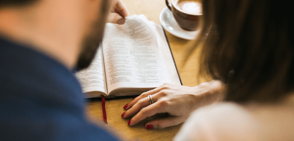 An adult couple study the Bible together, sharing their favorite scriptures while enjoying a cup of coffee. The man / husband reads aloud while pointing out a verse to his wife. Shot as viewed from behind and over their shoulders, the focus on the scriptures