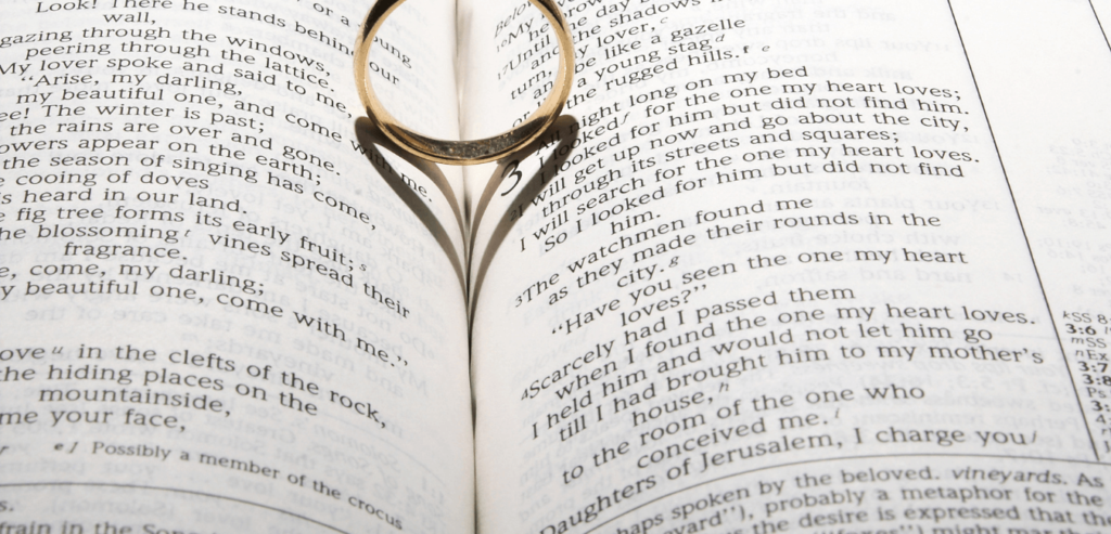 A wedding ring on a bible open to marriage scripture having a heart shaped shadow on it